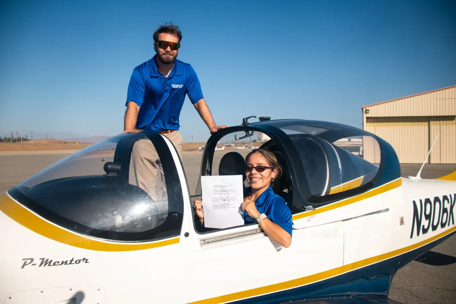 A student in the cockpit of a P-Mentor holding her new pilot certificate, with her instructor standing beside the canopy